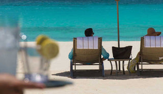 Image of couple sitting under a beach umbrella, about to get a drink from a waiter at the beach