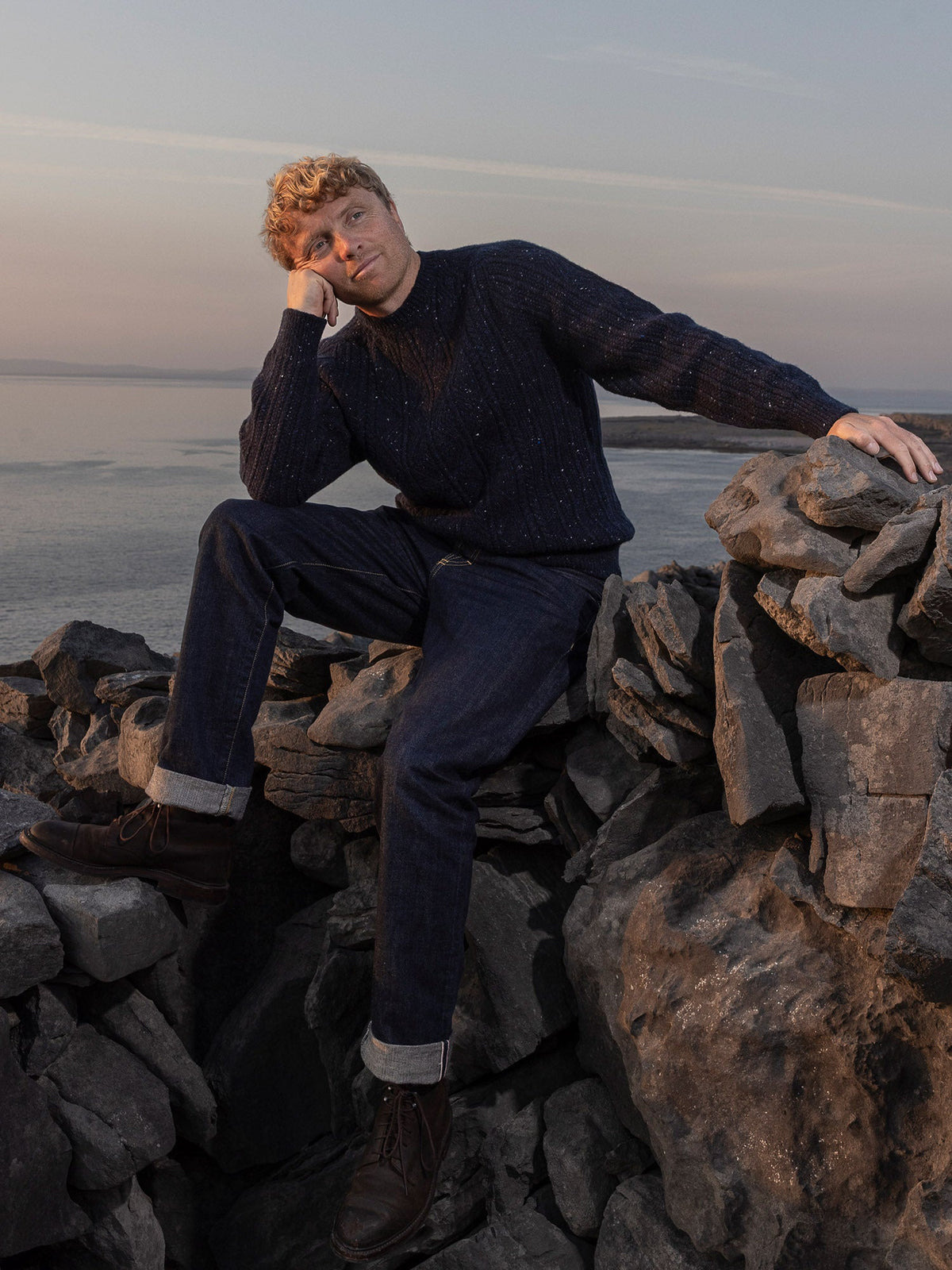 Inis Meáin Aran cable crewneck in navy, worn on a stone wall above the sea