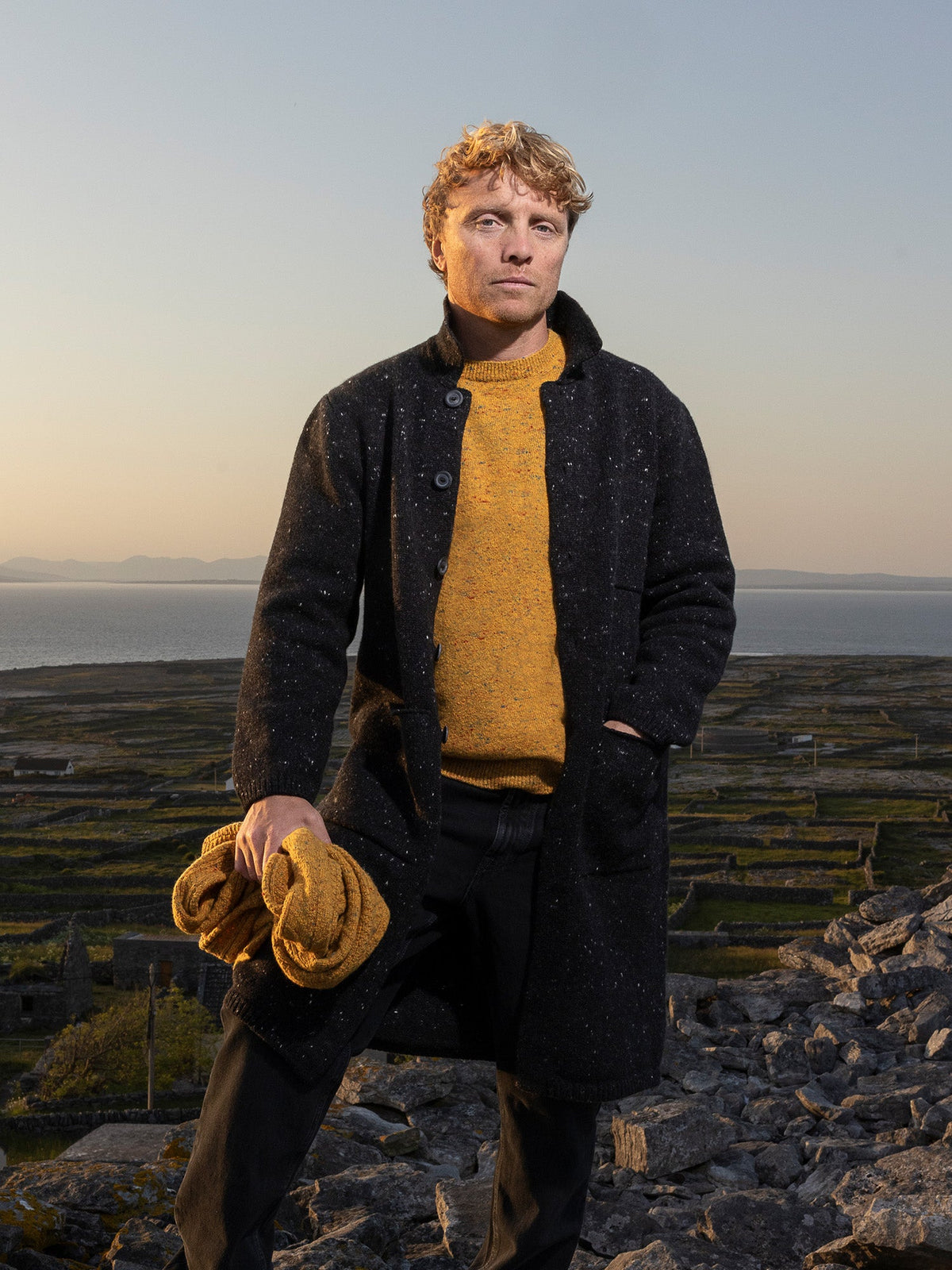 Inis Meáin Raftery coat in black fleck over a mustard crewneck, among stone walls at sunset
